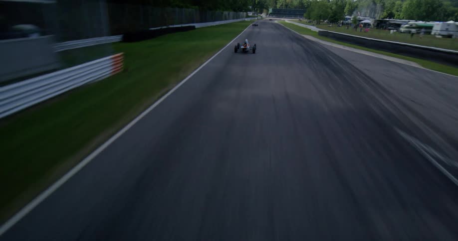 An aerial shot swoops down to follow a formula race car as it speeds around a track surrounded by lush greenery