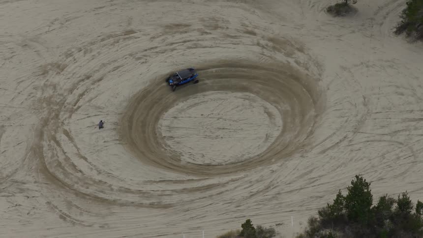 A high-angle aerial shot captures a blue UTV driving in circles, carving a large spiral pattern into the sand as a person watches