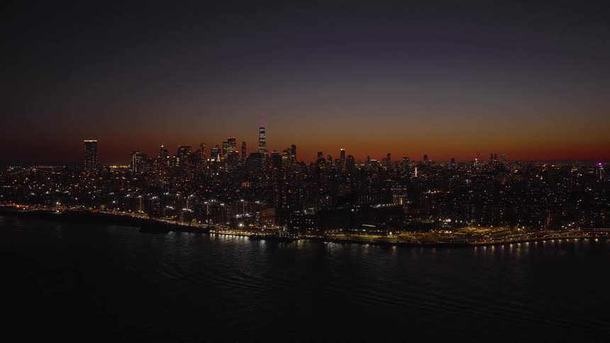 A tranquil aerial shot captures the sprawling, illuminated Lower Manhattan skyline against the colorful sky of dusk