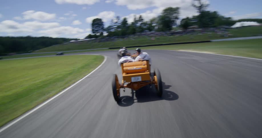 Dynamic camera follows a vintage orange race car as it speeds around a track during a classic car competition