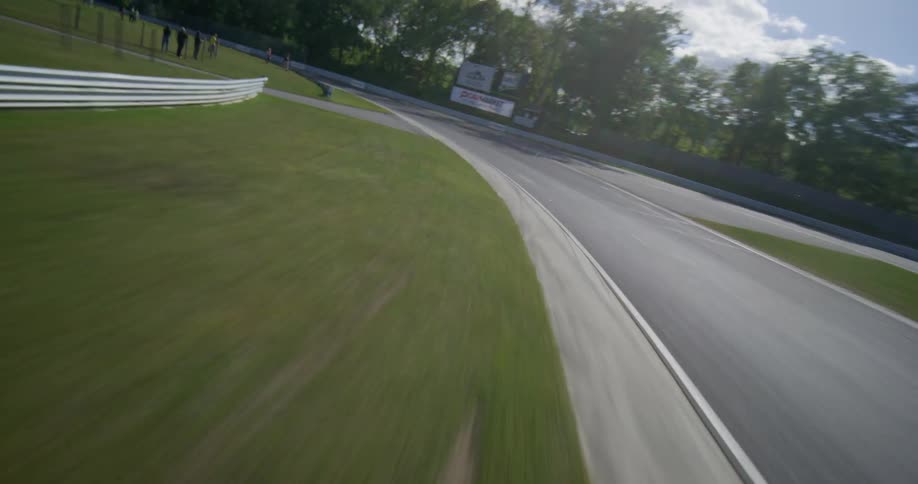 An exhilarating aerial shot transitions into a high-speed FPV drone flight, skimming the surface of a winding racetrack through a lush green landscape