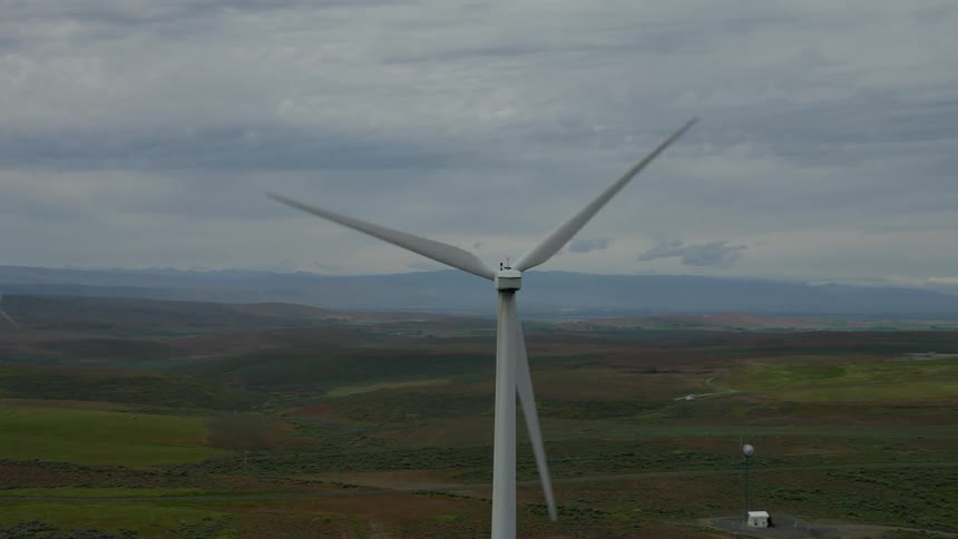 An aerial drone shot orbits a massive spinning wind turbine in a large wind farm set amongst rolling green hills under an overcast sky