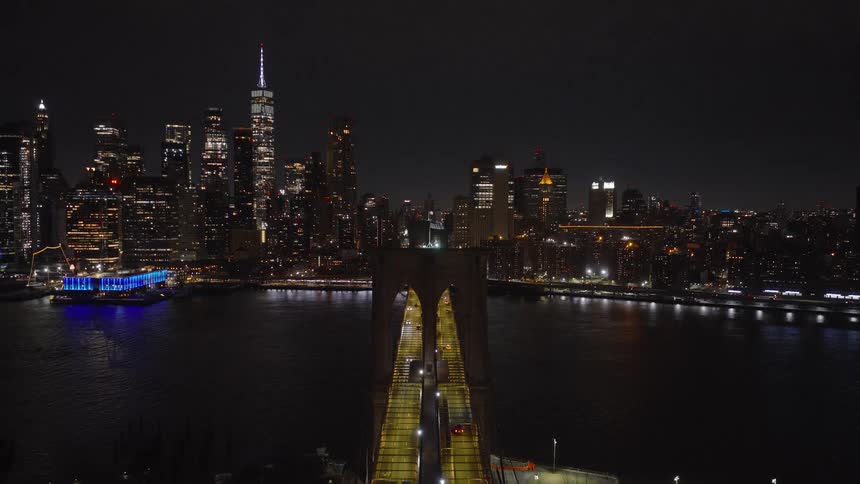 A dramatic aerial shot pulls back from the top of the Brooklyn Bridge at night, revealing the dazzling Manhattan skyline