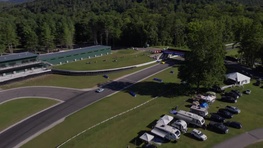 An aerial view of race cars competing on a winding race track through a lush green forest on a sunny day