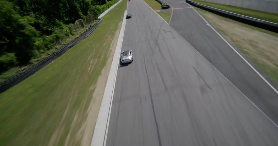 An exhilarating aerial view follows multiple race cars as they speed under a bridge and down the long straightaway of a scenic racetrack