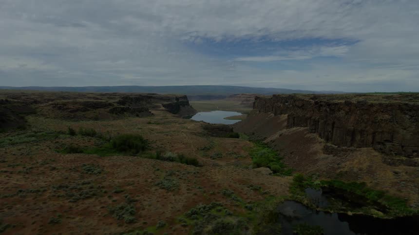 A slow aerial pullback reveals a vast, rugged canyon landscape with a hidden lake under a cloudy sky