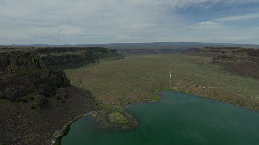 An epic aerial pullback shot reveals a tranquil green lake at the edge of a vast, arid valley surrounded by rugged cliffs