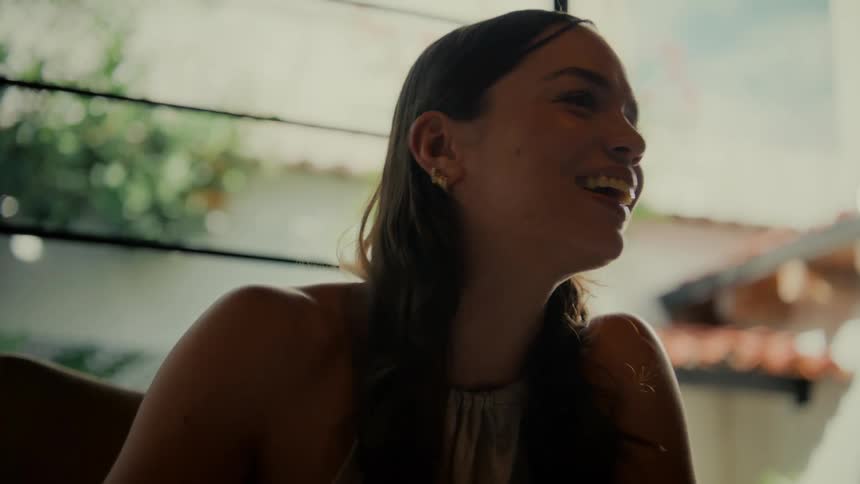 A thoughtful young woman sips her morning coffee on a sunlit patio, her mood shifting from pensive to a joyful laugh as she engages in conversation