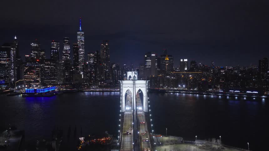 An aerial view pulls back from the Brooklyn Bridge traffic at night to reveal the iconic Manhattan skyline