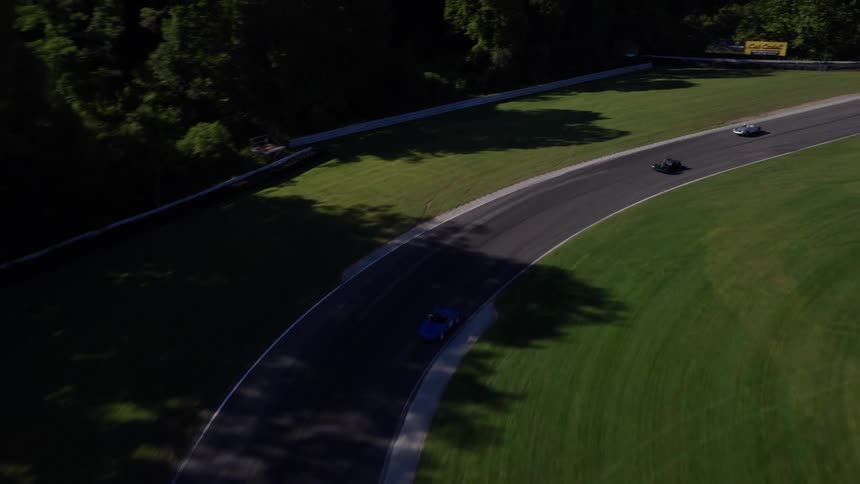 High-angle aerial view follows vintage race cars as they navigate a winding track through a verdant, hilly landscape during a sunny day race
