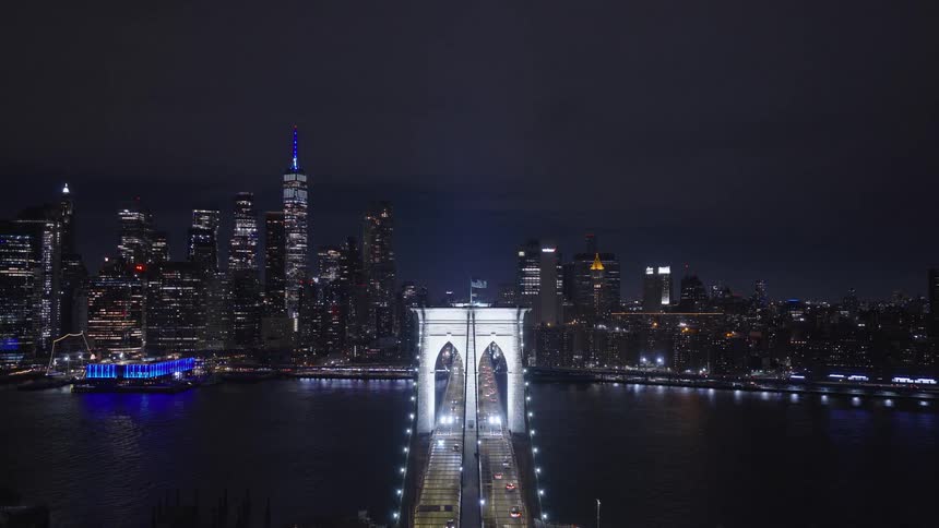 A slow aerial pullback reveals the iconic Brooklyn Bridge and the illuminated Manhattan skyline over the East River at night