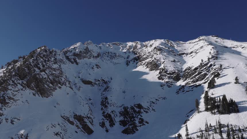 Majestic aerial pull-back shot reveals a vast, snow-covered mountain peak against a clear blue sky on a sunny winter day