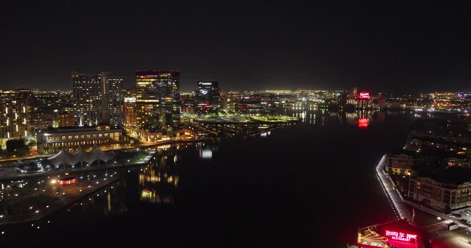 A slow-moving aerial shot captures the beautifully illuminated Baltimore Inner Harbor at night, with the glowing skyline reflecting on the water