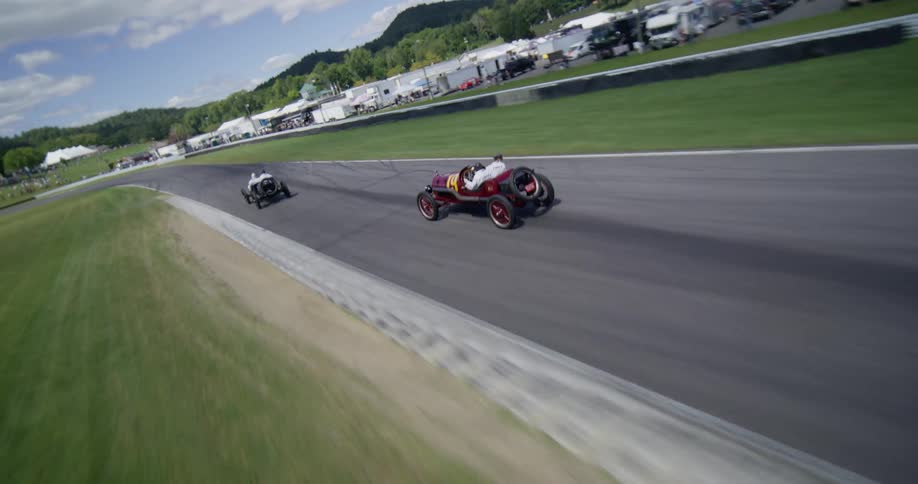 A dynamic follow and aerial shot captures a group of vintage race cars speeding around a modern racetrack on a sunny day