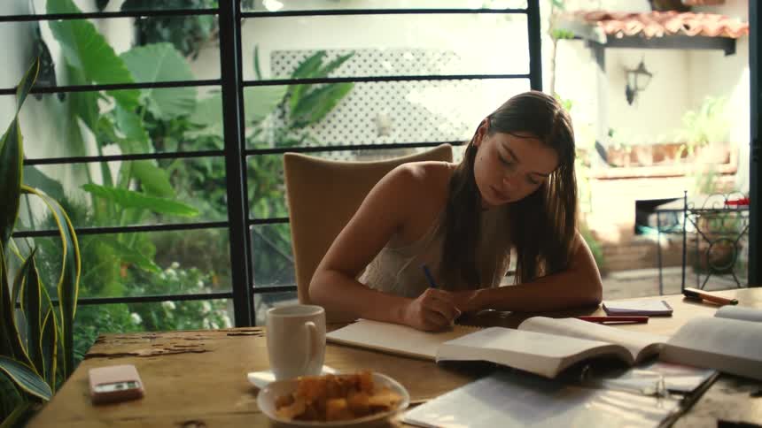 A focused female student studies diligently at a wooden desk by a large window overlooking a verdant garden