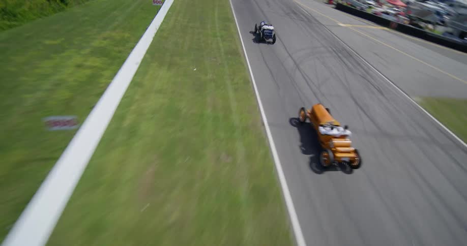 A high-speed FPV drone flies down a river and onto a racetrack to chase vintage race cars during a competition