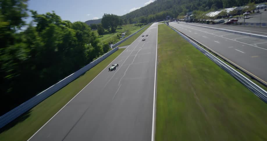 A dynamic aerial shot follows a group of vintage open-wheel race cars as they speed down the main straight at Lime Rock Park