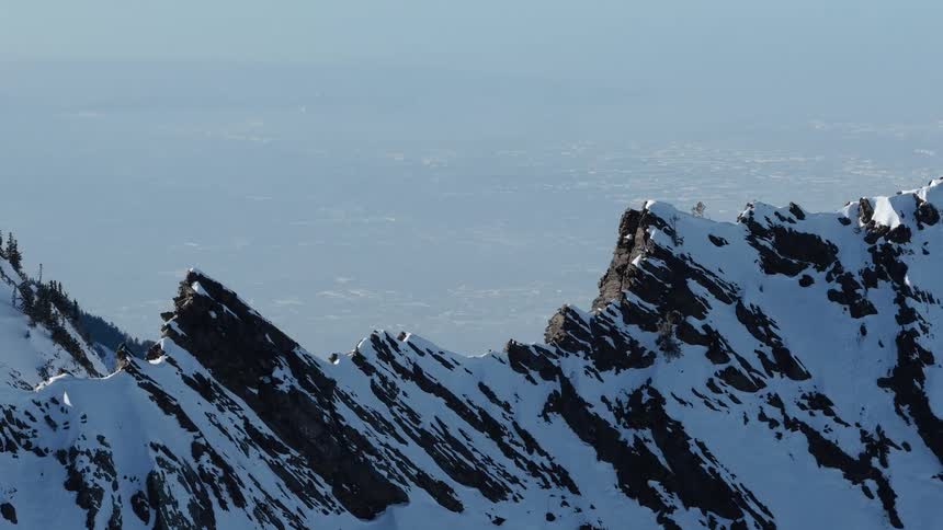 A rugged and snowy mountain ridgeline overlooks a hazy valley and distant cityscape