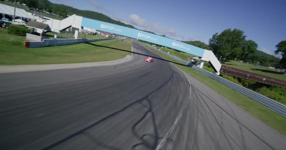 Dynamic aerial FPV drone footage follows colorful vintage race cars speeding around the scenic Lime Rock Park racetrack on a sunny day