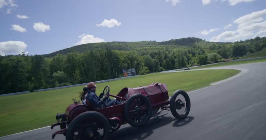 Dynamic tracking and aerial shots capture a classic red open-wheel race car speeding around a track during a vintage racing event