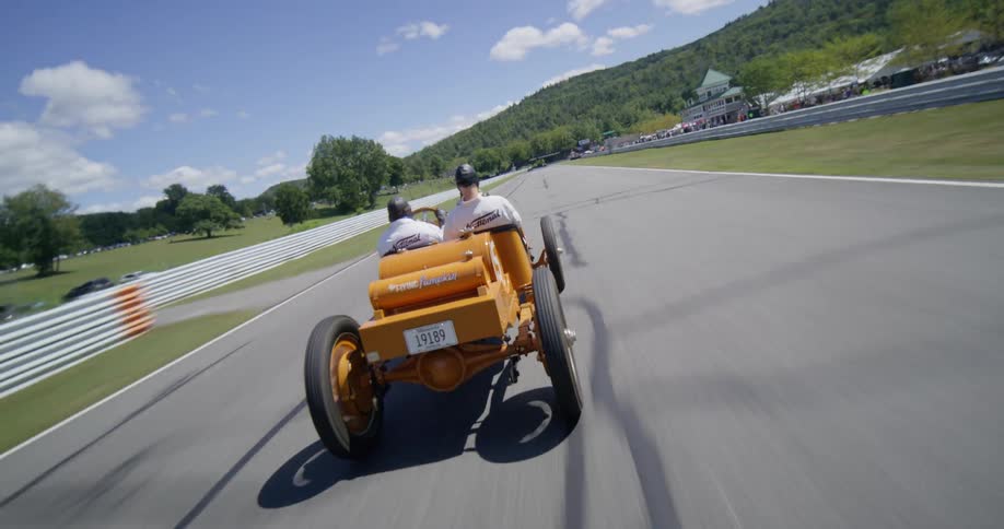 Aerial shot descends to follow a vintage orange race car speeding down a track during a sunny day event