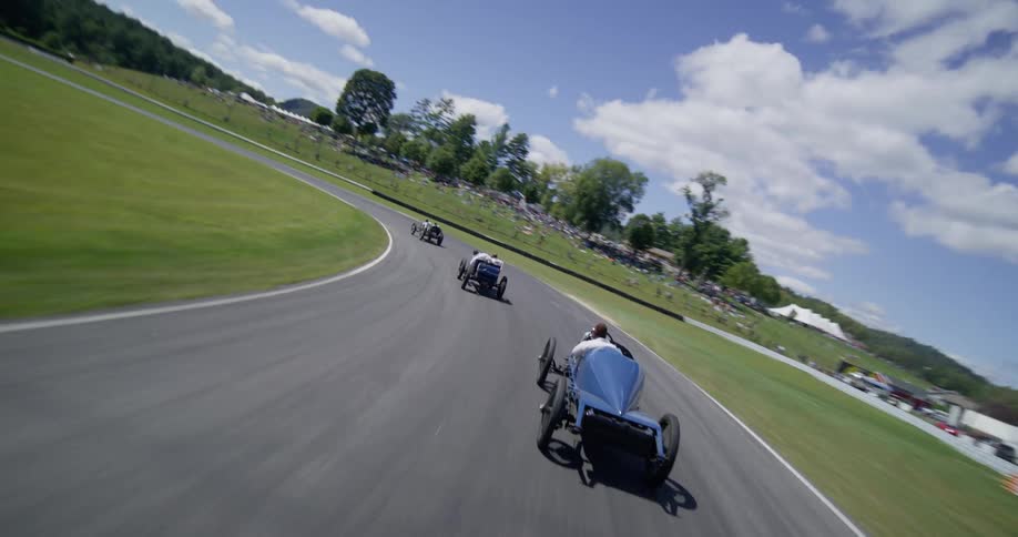 A dynamic FPV drone chases vintage open-wheel race cars around a scenic track on a sunny day