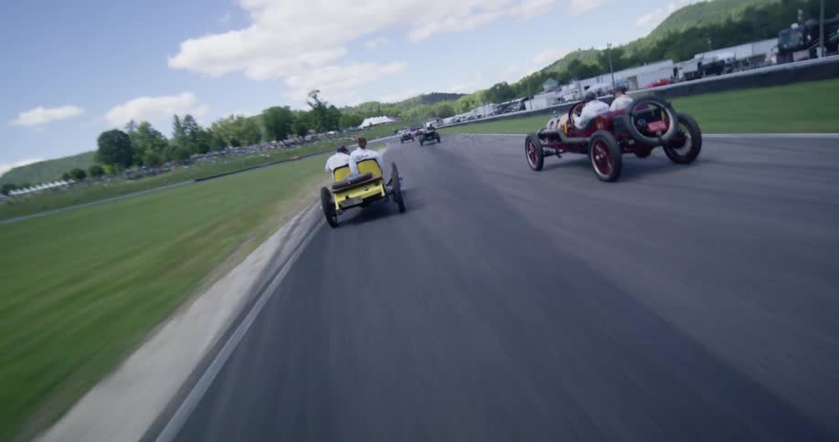 Dynamic aerial footage follows a fleet of classic early 20th-century race cars as they speed around a scenic, winding racetrack