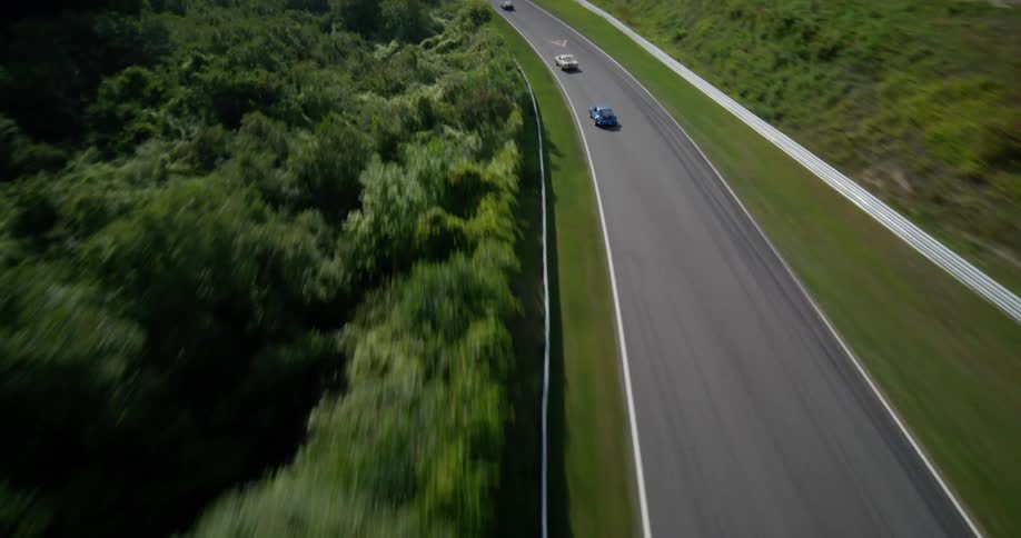 A thrilling FPV drone shot follows vintage sports cars as they speed around a scenic, winding race track