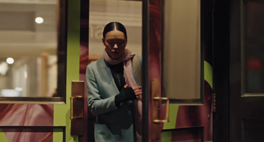 A stylishly dressed Asian woman walks out of a building's brightly decorated doorway into the night in slow motion