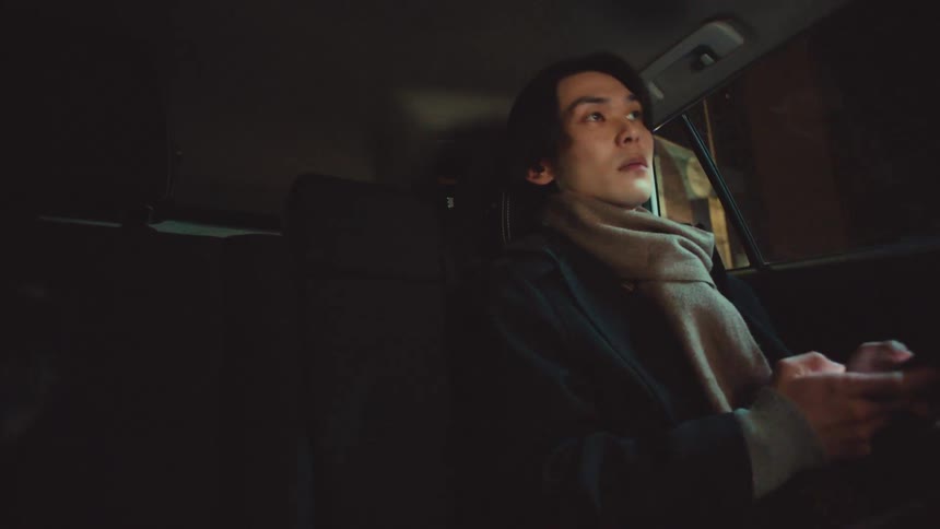A stylish young man uses his smartphone while riding in the back of a car at night, then looks out at the passing city lights