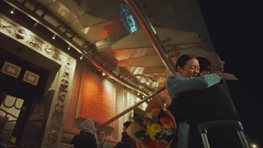 A happy woman greets her friend with a bouquet of flowers and a long, emotional hug upon her arrival at a grand venue at night