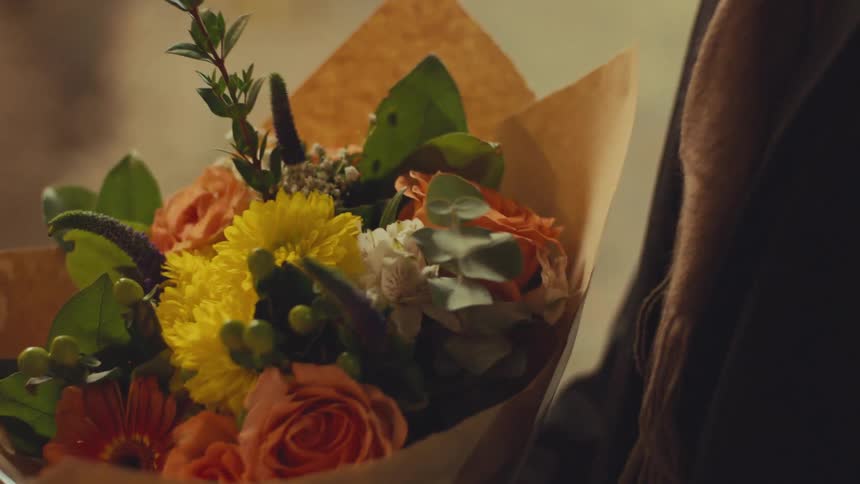 A person holds a vibrant bouquet of flowers wrapped in brown paper in a warmly lit, intimate setting