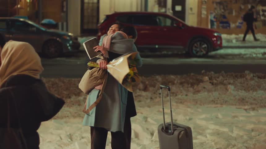 A happy man holding flowers greets his partner as she arrives with luggage, sharing a warm hug on a snowy city street at night