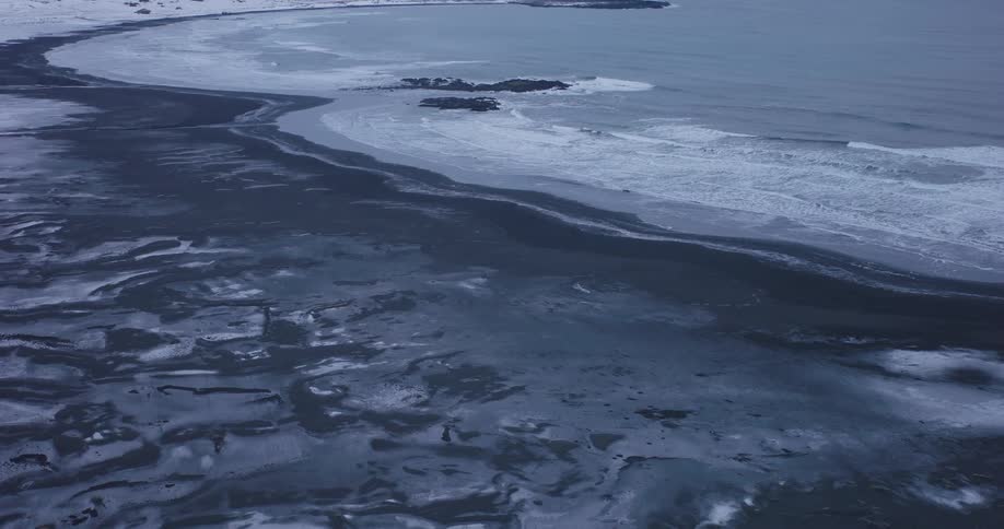 An expansive aerial shot slowly pulls back to reveal a vast, snow-dusted black sand beach meeting the cold ocean waves