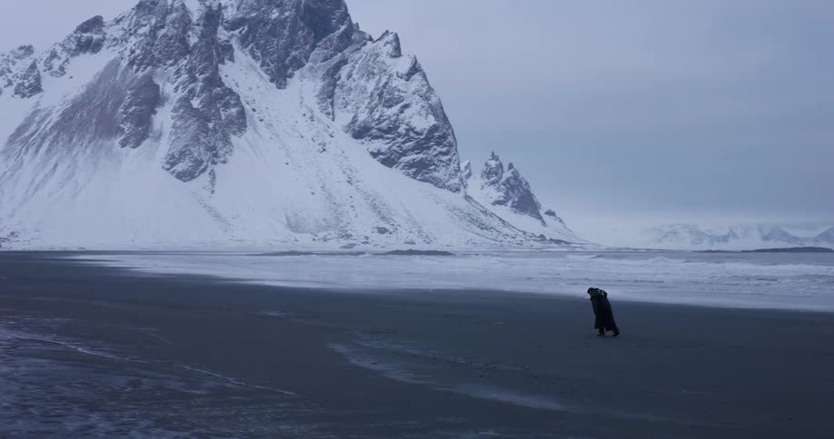 A mysterious cloaked figure walks alone on a vast black sand beach with immense, snowy mountains in the background