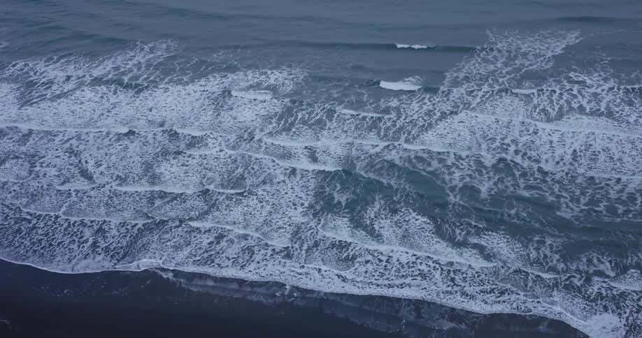 A mesmerizing slow-motion top-down aerial view of foamy ocean waves rolling onto a dark, sandy shoreline