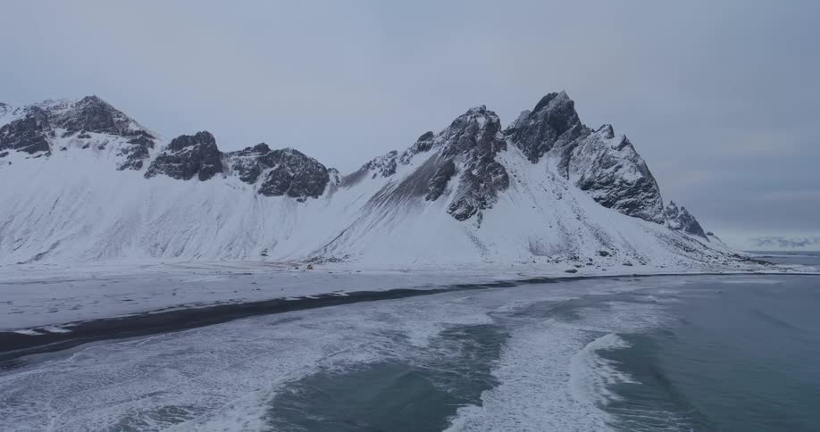 A majestic, snow-covered mountain range looms over a frozen black sand beach as ocean waves gently roll ashore in a serene winter landscape