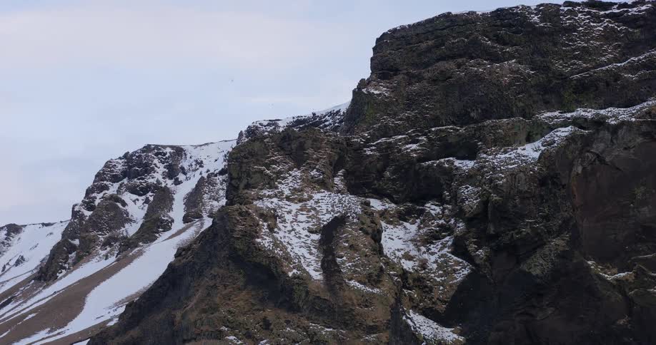 An aerial view pulls back from a craggy, snow-patched mountain revealing the vastness of the Icelandic highlands