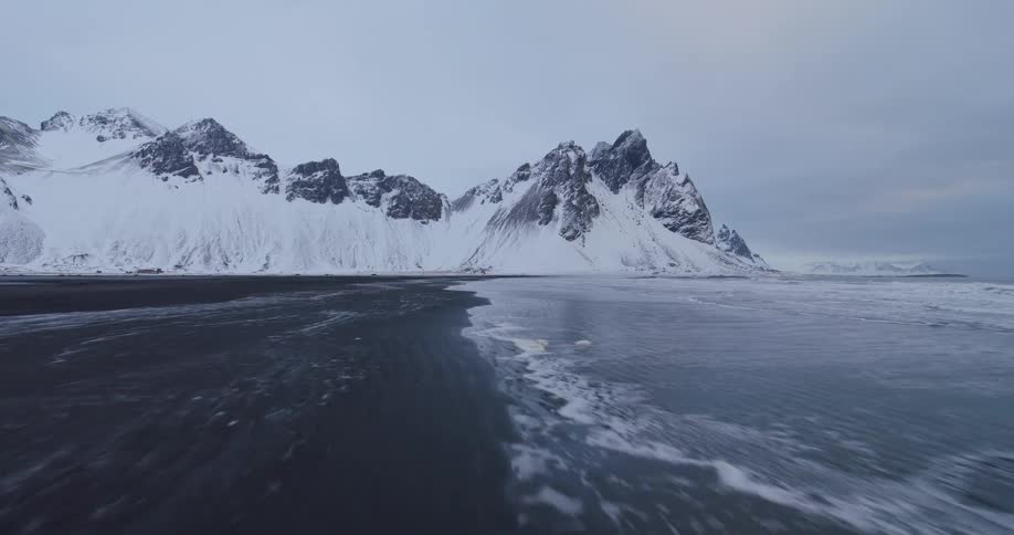 A dramatic low-angle aerial shot flies along the icy shoreline of a black sand beach with majestic snow-covered mountains in the background