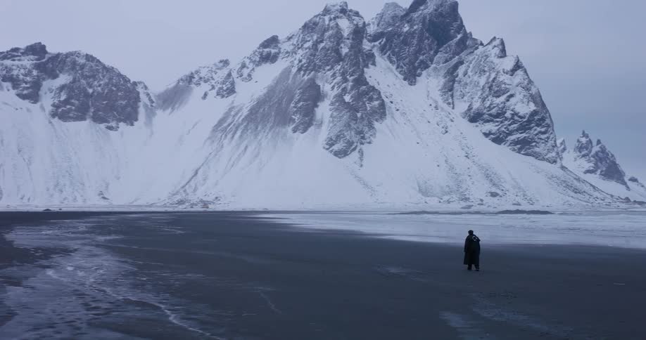 A lone figure walks across a vast, frozen black sand beach toward a majestic, snow-covered mountain range in Iceland