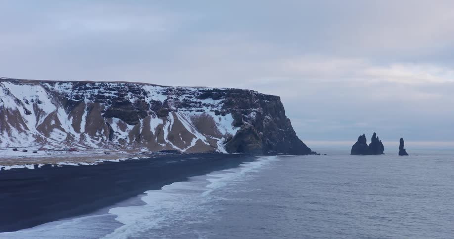 A majestic and desolate view of a snowy Icelandic coastline with a black sand beach and iconic sea stacks in the ocean