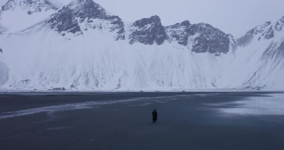 A lone figure walks across a vast, frozen beach, dwarfed by the immense snow-covered mountains of an epic winter landscape