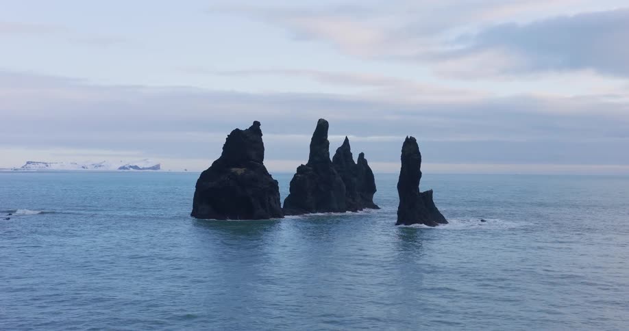 A slow, cinematic zoom-out reveals the iconic Reynisdrangar sea stacks standing in the calm ocean off the snowy coast of Iceland