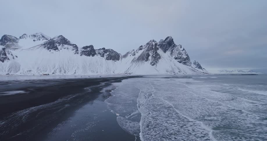 A slow aerial pullback reveals a majestic snow-covered mountain range reflecting on a black sand beach with icy waves
