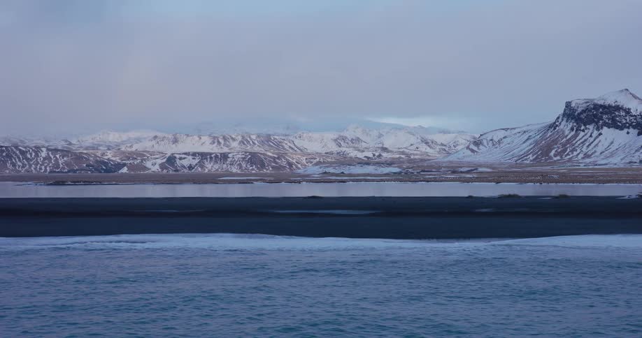 A tranquil winter scene shows gentle ocean waves on a black sand beach with a stunning backdrop of snow-covered mountains