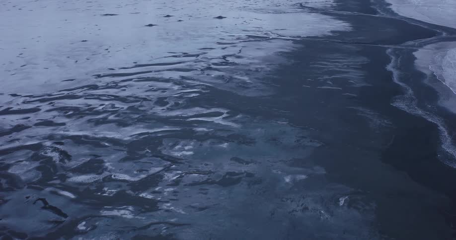 A stark and static aerial view shows a partially frozen body of water meeting an icy shore