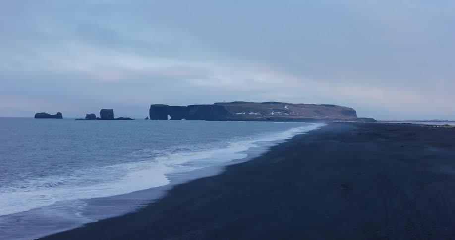 A dramatic aerial view pulls back to reveal the vast, moody landscape of an Icelandic black sand beach with waves rolling onto the shore