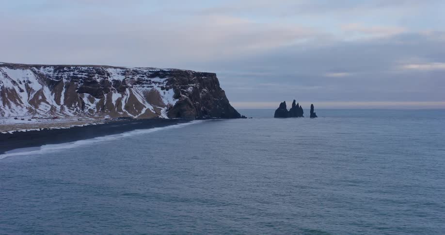 A serene, wide shot of the iconic Reynisdrangar sea stacks and snow-covered cliffs along the black sand beach of Reynisfjara in Iceland during winter