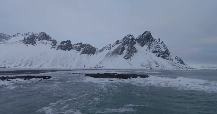 An epic slow-motion aerial flight over icy ocean waves towards the majestic snow-covered Vestrahorn mountain range in Iceland