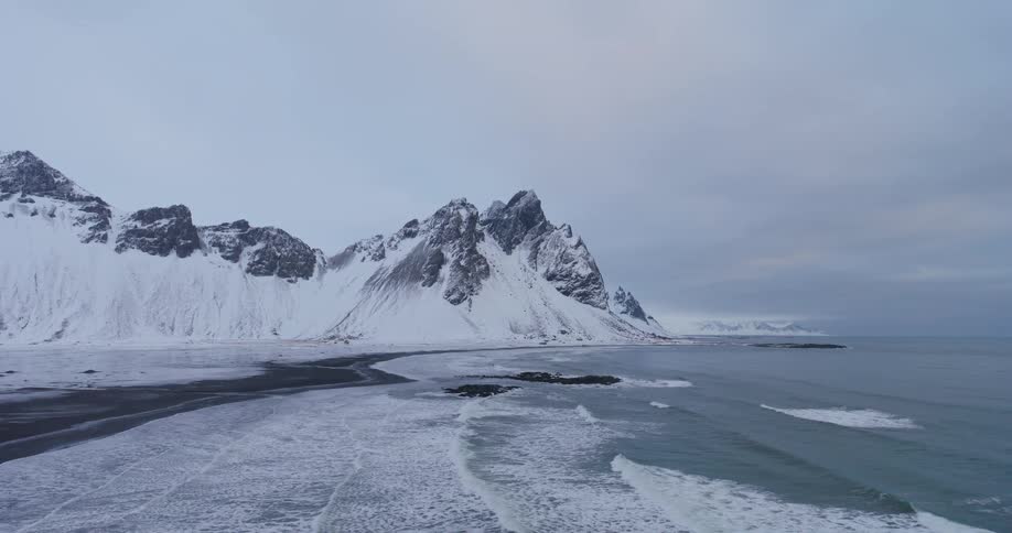 A majestic snow-covered mountain range in Iceland meets the icy ocean on a black sand beach under an overcast winter sky